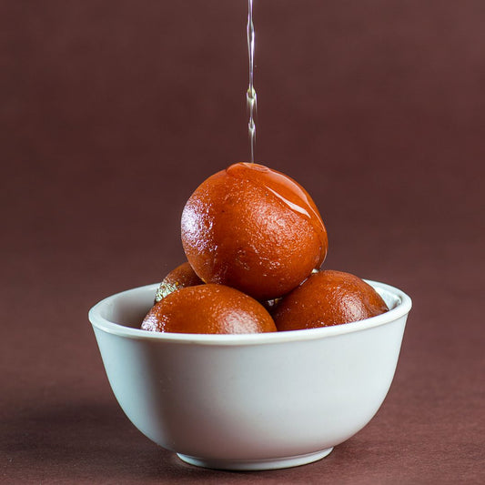 Gulab Jamun, un postre tradicional indio servido en un tazón blanco, con bolitas de masa frita empapadas en un jarabe dulce, con un chorro de jarabe que cae sobre ellas, presentado sobre un fondo marrón.